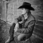 Black and white photograph of artist Chessney Sevier crouched on the ground wearing a black cowboy hat, jeans and cowboy boots